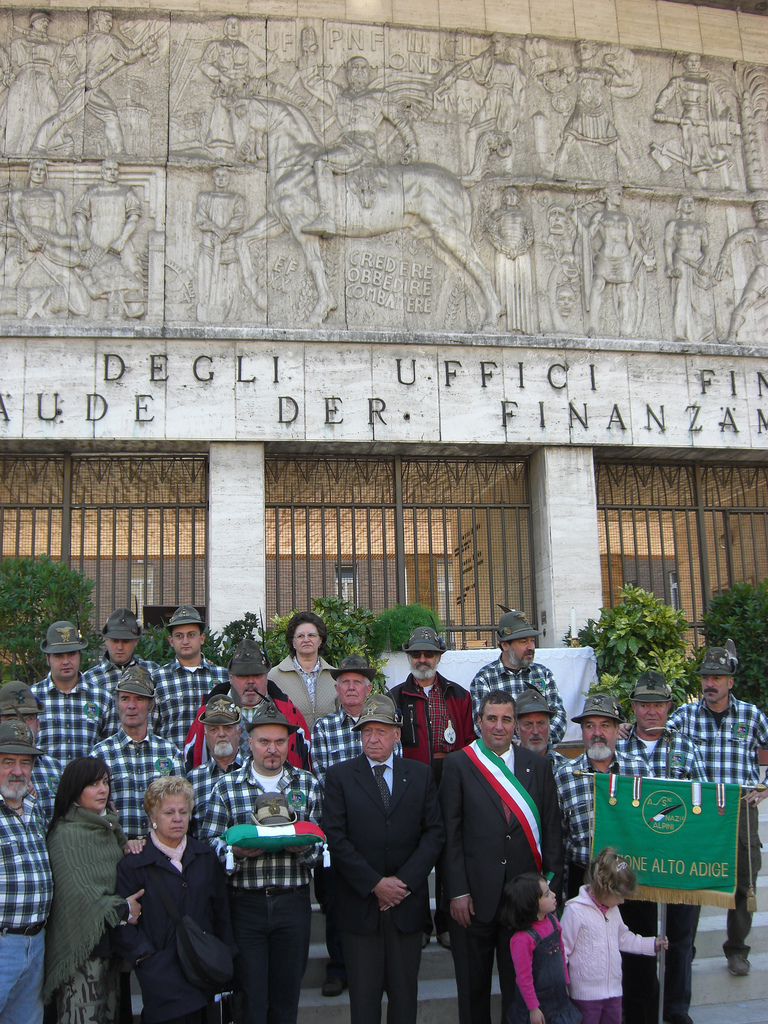 Alpini feiern vor dem Finanzamt in Bozen, welches mit einem Fries geschmückt ist, auf welchem der reitende „Duce“ Mussolini die Bürger dazu auffordert: „Glauben, gehorchen, kämpfen!“